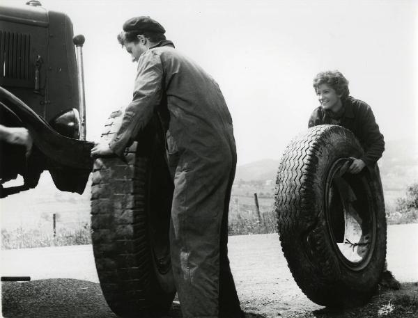 Scena del film "Esterina" - Regia Carlo Lizzani, 1959 - Vicino a un autoveicolo, un attore non identificato si appresta a cambiare un pneumatico mentre Carla Gravina avvicina un secondo pneumatico sorridendo.