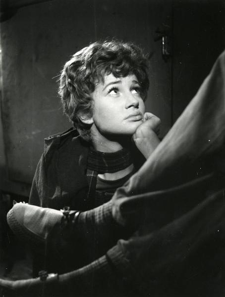 Scena del film "Esterina" - Regia Carlo Lizzani, 1959 - Mezza figura di Carla Gravina che, con la mano sinistra appoggiata sotto il mento, rivolge lo sguardo in alto a destra. In primo piano sono presenti le gambe di un attore non identificato.