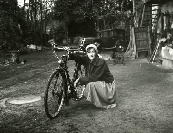 Scena del film "Esterina" - Regia Carlo Lizzani, 1959 - Carla Gravina, inginocchiata sorreggendo una bicicletta, rivolge lo sguardo verso l'obbiettivo.