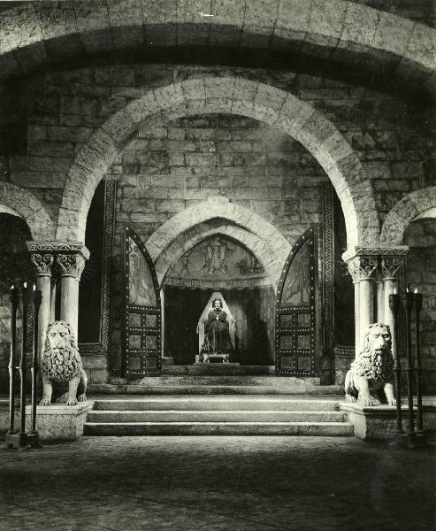 Scena del film "Ettore fieramosca" - Regia Alessandro Blasetti, 1938 - Elisa Cegani seduta su un trono di pietra. In primo piano, due leoni di pietra e quattro colonne portanti sorreggono una struttura ad arco.