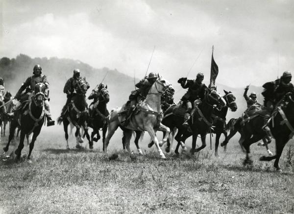 Scena del film "Ettore fieramosca" - Regia Alessandro Blasetti, 1938 - Scena di battaglia: una serie di cavalieri non identificati hanno le braccia e le spade alzate mentre sono al galoppo. Uno dei cavalieri al centro porta una bandiera.