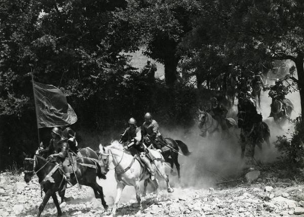 Scena del film "Ettore fieramosca" - Regia Alessandro Blasetti, 1938 - Numerosi attori non identificati a cavallo, sono in fila e si dirigono verso sinistra. Il primo attore a sinistra sorregge una bandiera aperta dal vento.