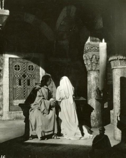 Scena del film "Ettore fieramosca" - Regia Alessandro Blasetti, 1938 - Un attore non identificato in abiti talari, seduto su una sedia a mani giunte, rivolge lo sguardo verso un'attrice non identificata di spalle, con vesti bianche, a destra.