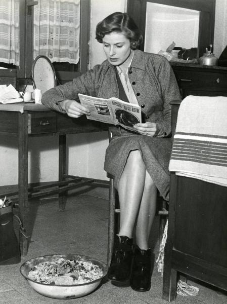 Scena del film "Europa 51" - Regia Roberto Rossellini, 1952 - Figura intera di Ingrid Bergman, seduta con un gomito appoggiato a una scrivania, intenta a leggere un libro intitolato "SCRITTO TRA GLI ASTRI".