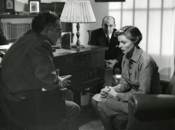 Scena del film "Europa 51" - Regia Roberto Rossellini, 1952 - Ingrid Bergman, seduta al centro con le mani giunte, rivolge lo sguardo verso il basso. Di fronte e dietro di lei, due attori non identificati seduti la guardano.