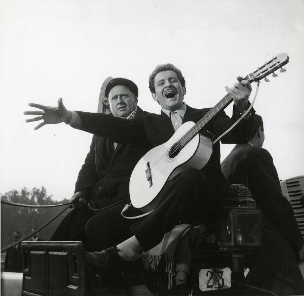 Scena del film "Europa di notte" - Regia Alessandro Blasetti, 1959 - Domenico Modugno, al centro e seduto su una carrozza con un attore non identificato, a sinistra, sorregge una chitarra e guarda verso l'obbiettivo.