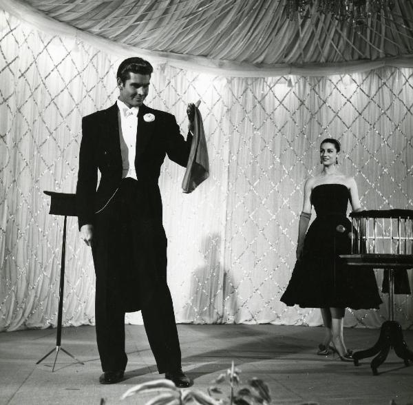 Scena del film "Europa di notte" - Regia Alessandro Blasetti, 1959 - Channing Pollock, in primo piano in frac, regge con la mano sinistra una stoffa e la osserva sorridente. A destra, in abiti eleganti, Alba Arnova lo osserva.