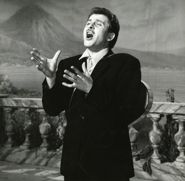 Scena del film "Europa di notte" - Regia Alessandro Blasetti, 1959 - Mezza figura di Domenico Modugno che canta, con una chitarra appoggiata alla schiena, alzando le mani vicino al torace.