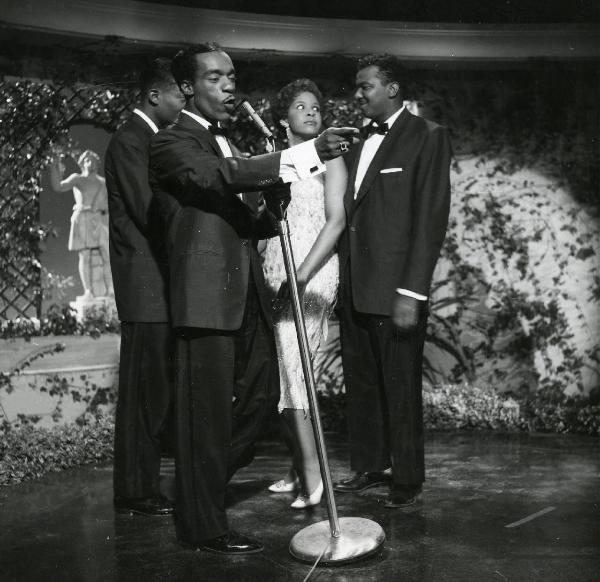 Scena del film "Europa di notte" - Regia Alessandro Blasetti, 1959 - In secondo piano, attori non identificati si osservano. In primo piano, un attore non identificato canta e indica con la mano destra davanti a sé.