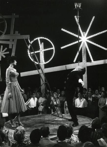 Scena del film "Europa di notte" - Regia Alessandro Blasetti, 1959 - Su un piccolo palco: Alba Arnova osserva un attore non identificato che, inclinato all'indietro, regge sulla bocca un vassoio con dei bicchieri sostenuto da due spade.