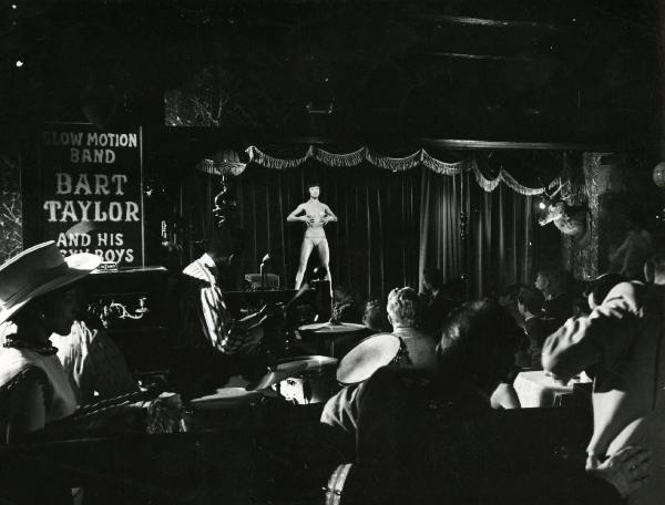 Scena del film "Europa di notte" - Regia Alessandro Blasetti, 1959 - In primo piano un'orchestra suona e degli attori non identificati osservano in direzione di un palcoscenico. Qui, Lady Phu Qui Cho, seminuda, si porta le mani vicino al petto.