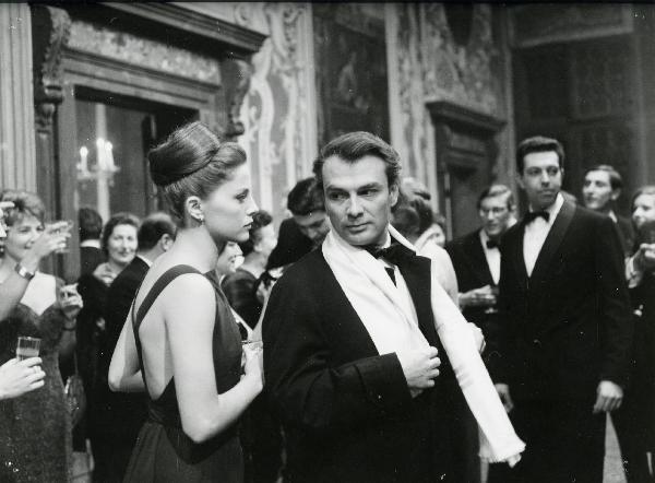 Scena del film "Eva" - Regia Joseph Losey, 1962 - In primo piano, in abiti eleganti, Virna Lisi, a sinistra, e Giorgio Albertazzi, a destra, si osservano. Alle loro spalle, numerosi attori e attrici non identificati.