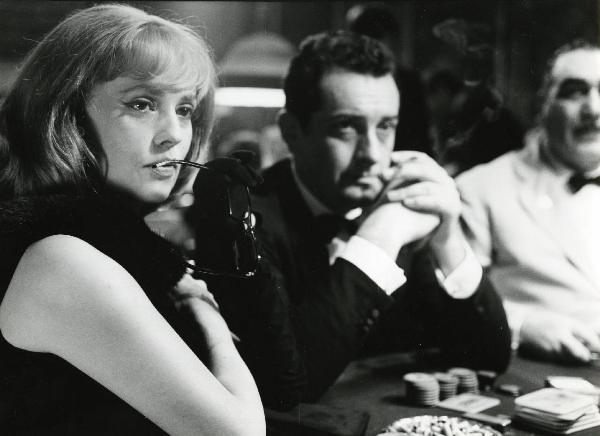 Scena del film "Eva" - Regia Joseph Losey, 1962 - In primo piano Jeanne Moreau, portandosi gli occhiali alla bocca, guarda davanti a sé, come Riccardo Garrone, al centro, e un attore non identificato in secondo piano.