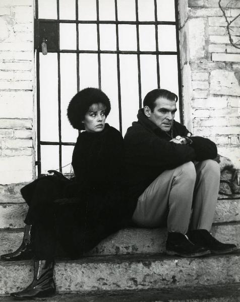 Scena del film "Eva" - Regia Joseph Losey, 1962 - Davanti a un cancello in metallo, seduti su dei gradini, Jeanne Moreau osserva verso sinistra in direzione di Stanley Baker che rivolge lo sguardo dritto davanti a sé.