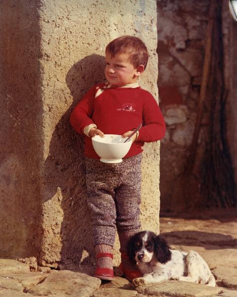 Scena del film "E venne un uomo" - Regia Ermanno Olmi, 1965 - Figura intera di un giovane attore, in piedi, appoggiato a un muro con una scodella in mano con un piccolo cane appoggiato alla sua gamba sinistra.