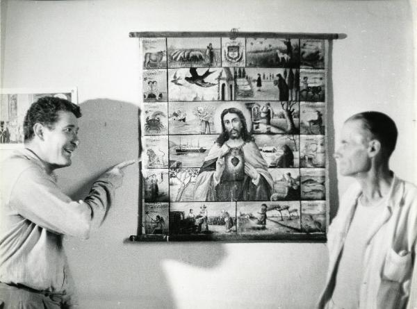 Scena del film "E venne un uomo" - Regia Ermanno Olmi, 1965 - Un attore non identificato, a sinistra, mostra con la mano destra un manifesto votivo che riporta l'immagine del Cristo e dei segni zodiacali ad un altro attore non identificato, a destra.