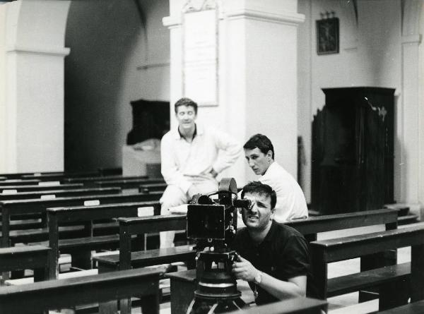 Sul set del film "E venne un uomo" - Regia Ermanno Olmi, 1965 - In una chiesa: un operatore in primo piano è intento a riprendere davanti a sé. Alle sue spalle altri due operatori non identificati osservano la scena.