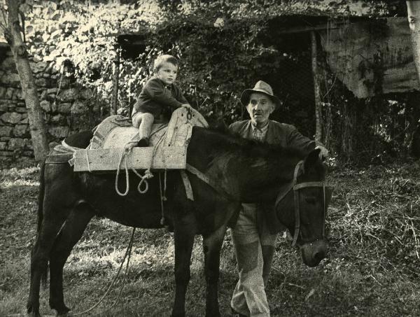 Scena del film "E venne un uomo" - Regia Ermanno Olmi, 1965 - Un giovane attore è seduto su una sella di legno su un asino. Un altro attore non identificato, posando la mano sinistra sul capo dell'animale, guarda davanti a sé.