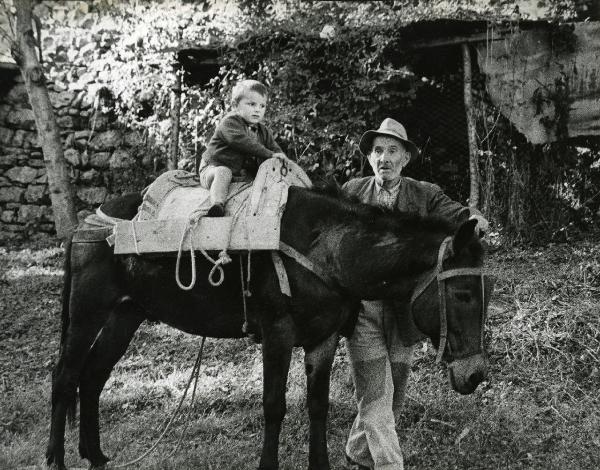 Scena del film "E venne un uomo" - Regia Ermanno Olmi, 1965 - Un giovane attore è seduto su una sella di legno su un asino. Un altro attore non identificato, posando la mano sinistra sul capo dell'animale, guarda davanti a sé.