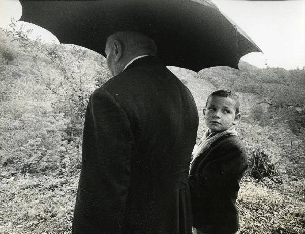 Scena del film "E venne un uomo" - Regia Ermanno Olmi, 1965 - Un attore non identificato di spalle, con un ombrello aperto, tiene per mano un bambino che guarda verso l'obbiettivo.