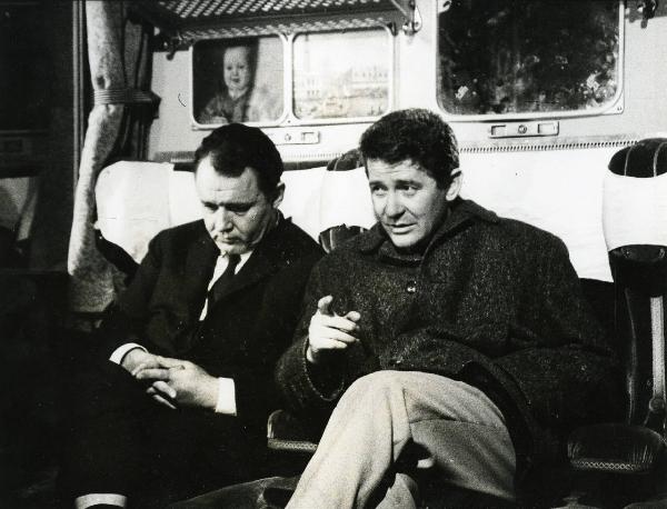 Scena del film "E venne un uomo" - Regia Ermanno Olmi, 1965 - Due attori non identificati seduti sul treno: l'attore a sinistra guarda verso il basso, mentre quello a destra con un dito indica davanti a sé mentre tiene l'altra mano in tasca.