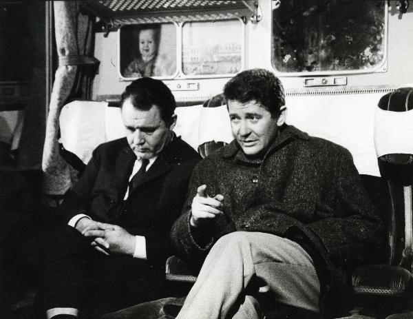 Scena del film "E venne un uomo" - Regia Ermanno Olmi, 1965 - Due attori non identificati seduti sul treno: l'attore a sinistra guarda verso il basso, mentre quello a destra con un dito indica davanti a sé mentre tiene l'altra mano in tasca.