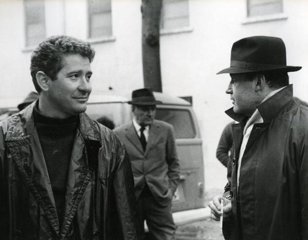 Scena del film "E venne un uomo" - Regia Ermanno Olmi, 1965 - In primo piano a sinistra, un attore non identificato guarda verso destra mentre un secondo attore non identificato, quasi completamente voltato di spalle, tiene in mano una sigaretta.