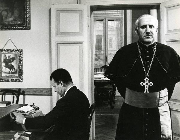 Scena del film "E venne un uomo" - Regia Ermanno Olmi, 1965 - Un attore non identificato seduto a sinistra digita su una macchina da scrivere. A destra, un attore non identificato in veste chiericale guarda davanti a sé con le mani dietro la schiena.