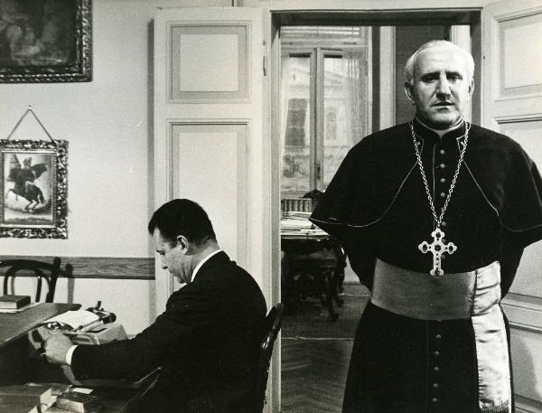 Scena del film "E venne un uomo" - Regia Ermanno Olmi, 1965 - Un attore non identificato seduto a sinistra digita su una macchina da scrivere. A destra, un attore non identificato in veste chiericale guarda davanti a sé con le mani dietro la schiena.