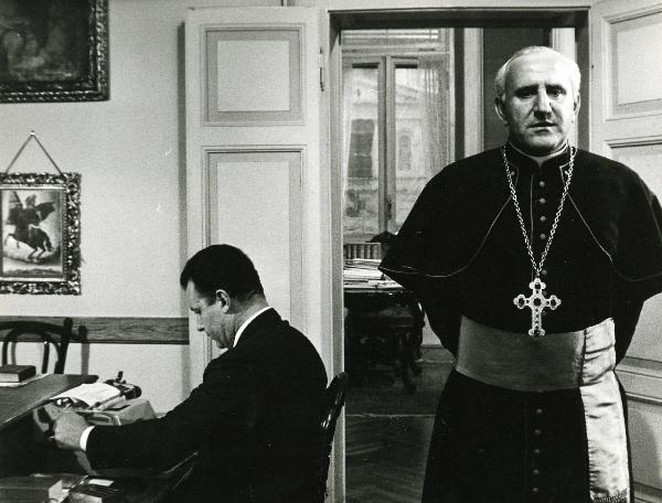 Scena del film "E venne un uomo" - Regia Ermanno Olmi, 1965 - Un attore non identificato seduto a sinistra digita su una macchina da scrivere. A destra, un attore non identificato in veste chiericale guarda davanti a sé con le mani dietro la schiena.