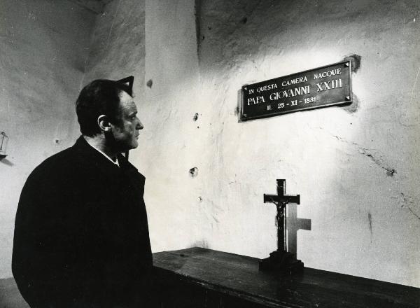 Scena del film "E venne un uomo" - Regia Ermanno Olmi, 1965 - A sinistra, Rod Steiger, rivolto verso destra, fissa la targa dedicata a Papa Giovanni XXIII. Sul mobile di legno, un piccolo crocifisso.