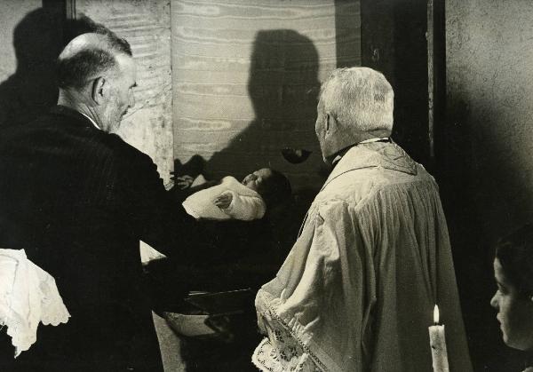 Scena del film "E venne un uomo" - Regia Ermanno Olmi, 1965 - Due attori non identificati di spalle battezzano un bambino. Nell'angolo a destra, spunta il viso di un chierichetto che tiene in mano una candela accesa.