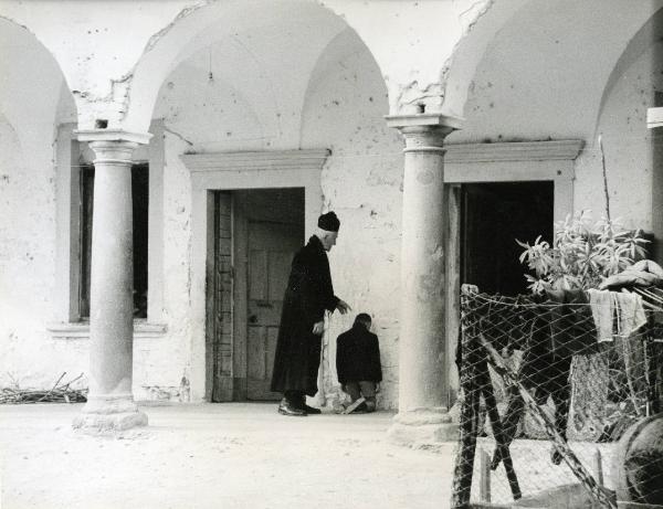 Scena del film "E venne un uomo" - Regia Ermanno Olmi, 1965 - Sotto a un portico ci sono due attori non identificati. Il bambino è in ginocchio con il viso rivolto verso il muro mentre l'uomo in vesti di prete lo indica e si rivolge a lui.