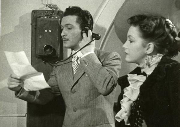 Scena del film "La fabbrica dell'imprevisto" - Regia Jacopo Comin, 1942 - Maurizio D'Ancora parla al telefono tenendo nella mano sinistra un foglio di carta. A destra, Vera Bergman, con indosso un cappello, lo osserva.