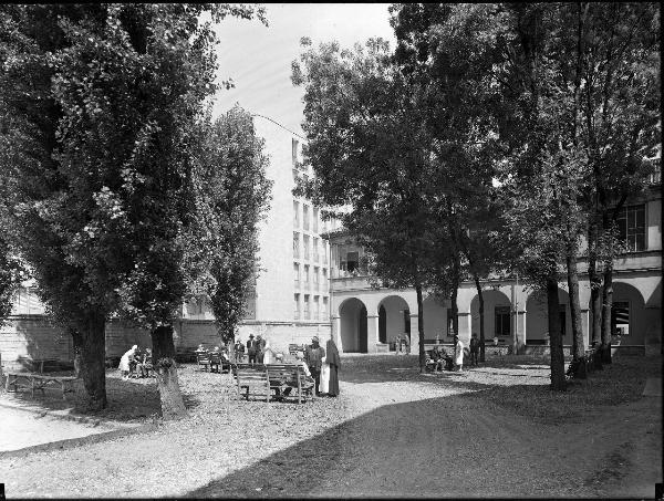 Pavia - Viale Matteotti 63 - Casa di riposo «F. Pertusati» - Cortile interno - Gruppi di uomini anziani seduti sulle panchine, accanto sono visibili due infermiere e due suore