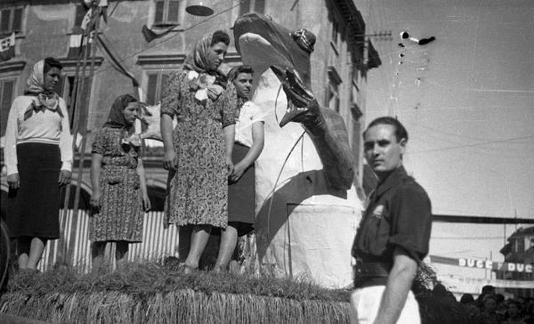 Garlasco - Festa delle mondine - Carro allegorico con quattro donne con abiti tradizionali e una rana gigante in cartapesta - Giovane uomo accanto al carro fissa l'obiettivo - Scorcio di palazzo sullo sfondo