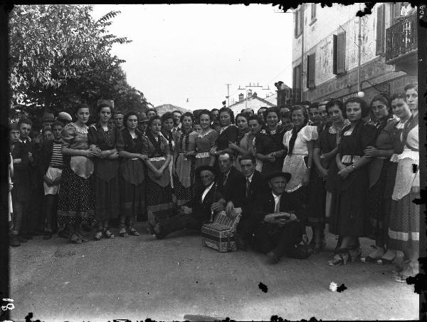 Ritratto di gruppo - Donne in abiti tradizionali e gruppo di uomini seduti a terra con strumento musicale a fiato e fisarmonica - Salice Terme - IX Festa nazionale dell'uva