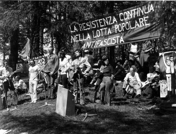 Festa Partigiana. Spettacolo di canzoni sulla Resistenza. Gruppo musicale all'aperto. Striscione con scritta "La Resistenza continua nella lotta popolare antifascista".