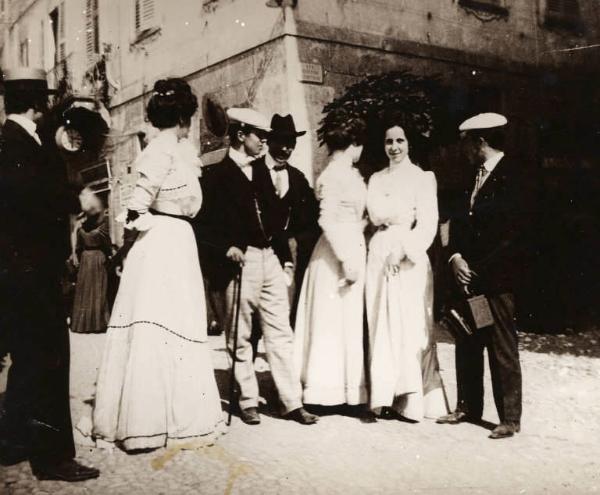 Personaggi davanti al palazzo municipale. Uomini e donne a passeggio. Uno di loro con macchina fotografica