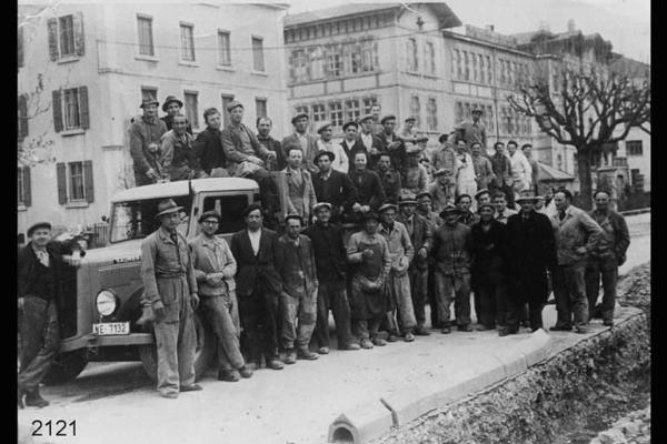 Emigranti valdimagnini e operai locali. Col basco, accanto alla portiera del camion, Francesco Facchinetti. Posa in esterno attorno e sopra un camion, lungo strada urbana.