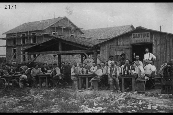 Emigranti. Cantiere a Besançon. Posa in esterno: gli uomini sono seduti ai tavoli con bottiglie; dietro a loro una capanna con un cartello: "Cantine - Paganelli". Festa per l'ultimazione del tetto.