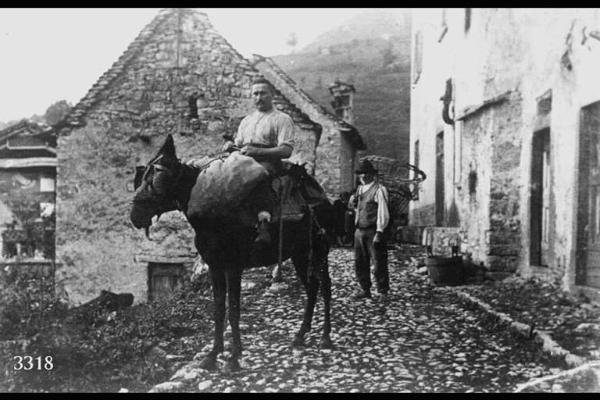 Uomo con sacco in groppa ad un mulo e contadino con gerla. Strada di paese acciottolata ed edifici rustici.