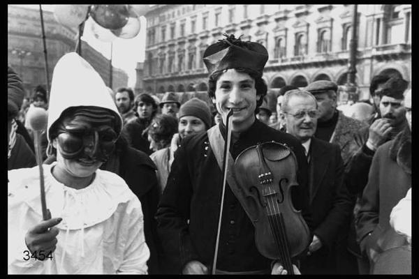 Carnevale ambrosiano. Piazza duomo: in primo piano due persone mascherate da Pulcinella e da prete che suona il violino.