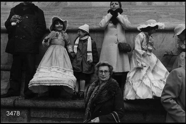 Carnevale ambrosiano. Piazza del Duomo: bambini in costume e adulti appoggiati al muro della Cattedrale.