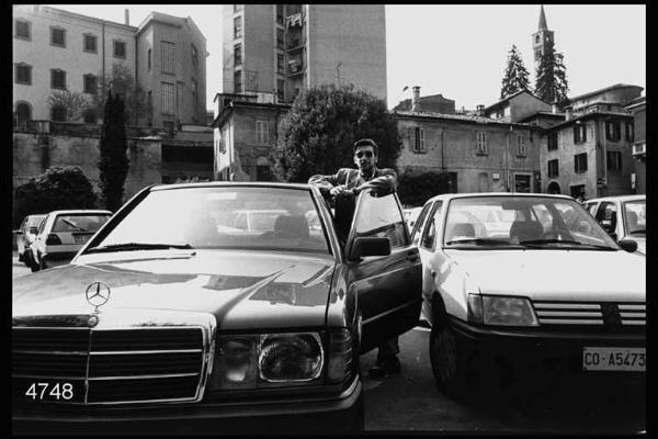 Parcheggio comunale. Giovane al fianco di una Mercedes.