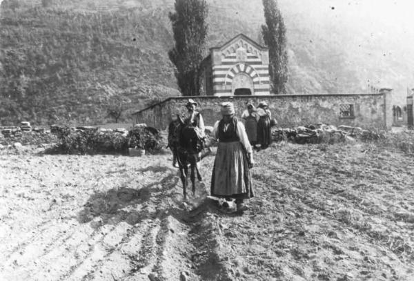 Uomo e donna che arano un campo. Sullo sfondo un cimitero.