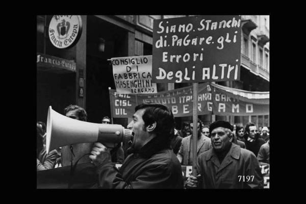 Manifestazione sindacale. In primo piano, lavoratore che urla nel megafono. Sullo sfondo cartelli e striscioni di consigli di fabbrica.