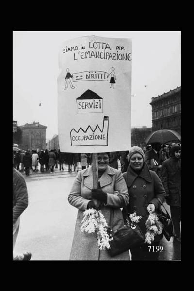 Manifestazione per la giornata internazionale della donna. In primo piano coppia di donne con in mano mazzetti di mimosa e un cartello.