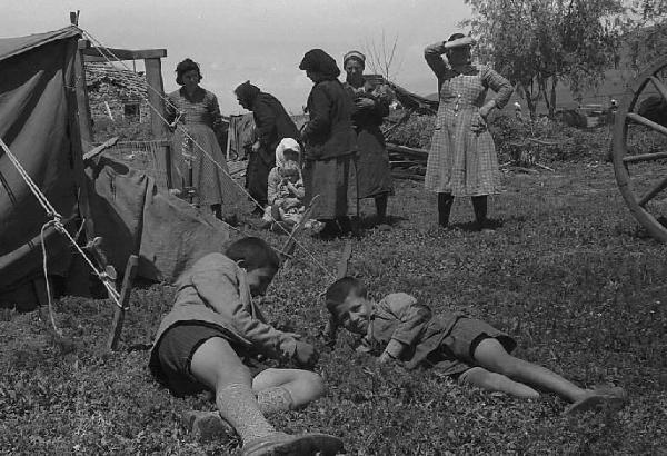 Località non identificata.Terremoto. Bambini coricati sull'erba. Sullo sfondo un gruppo di donne e le macerie provocate dal sisma