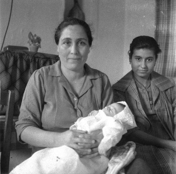 Melissa (Crotone) - Donna con neonato in abito da battesimo e altra donna in un interno di casa